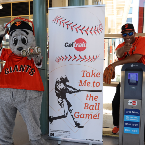 Lou Seal and person posing next Caltrain Banner and ticket machine